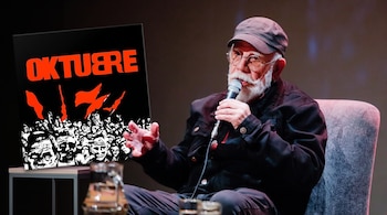 Un hombre con barba blanca, gafas y gorra habla ante un micrófono junto a la portada ilustrada del disco Oktubre en tonos negro, blanco y rojo.