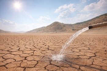 Un chorro de agua cae desde una manguera hacia el suelo agrietado y seco de un paisaje desértico, bajo un cielo azul con nubes dispersas y un sol intenso.