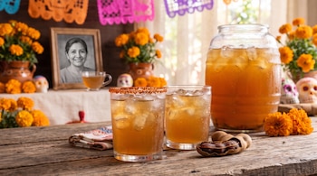 Dos vasos con agua fresca y hielo, un vitrolero, vainas de tamarindo, flores de cempasúchil y un retrato enmarcado sobre una mesa de madera.
