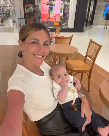 Mujer con camiseta blanca y pantalón negro sostiene a un bebé con polo blanco. El bebé tiene un chupete. Se ven mesas, sillas de madera y tiendas de ropa al fondo