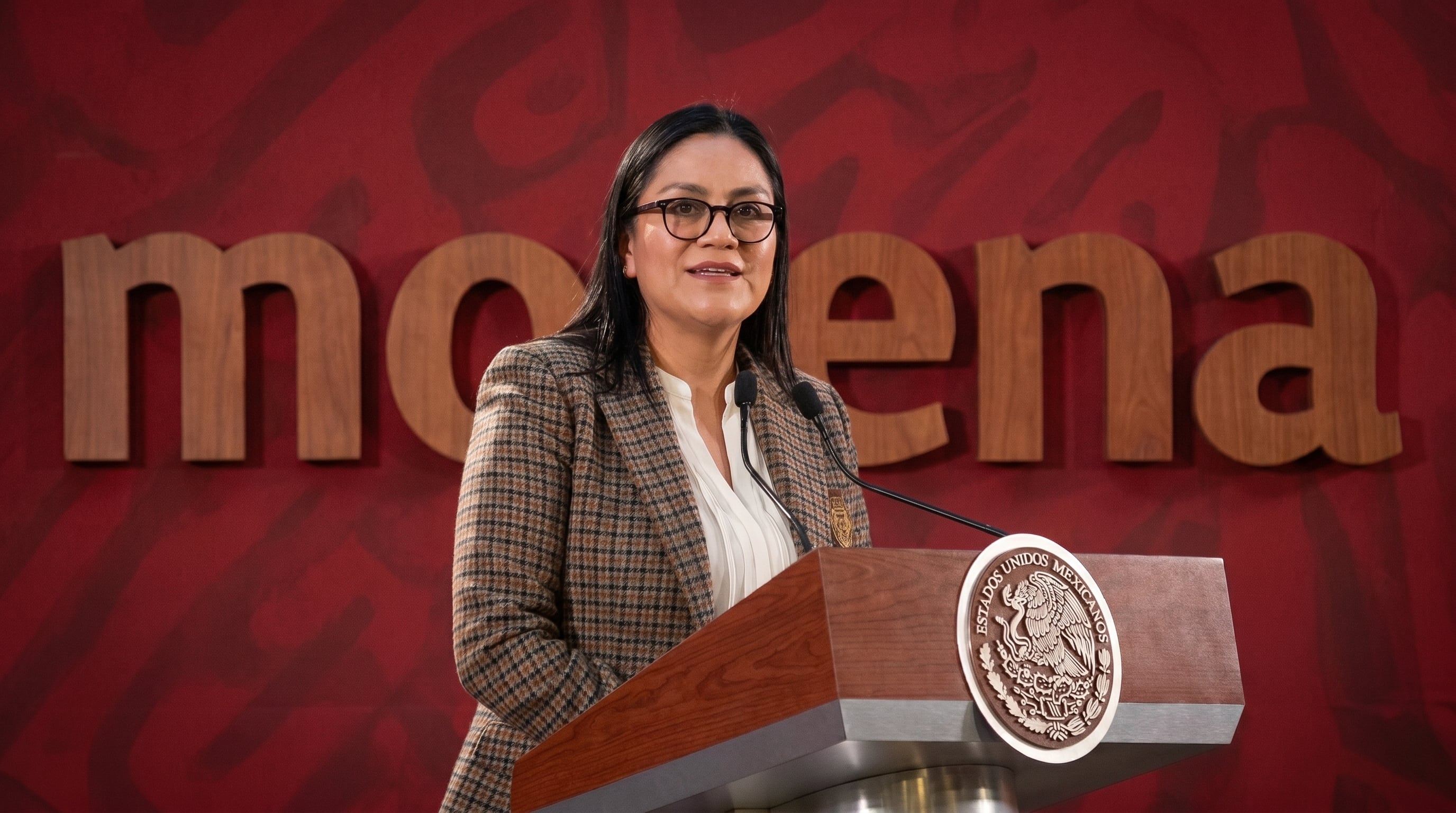 Una mujer sonríe y habla desde un podio de madera con el escudo nacional mexicano, frente a un muro rojo con el logo