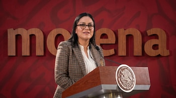 Mujer de cabello oscuro y gafas, vestida con blazer a cuadros marrones y camisa blanca, sonriendo y hablando ante un podio. Detrás, un muro rojo con el logo 'morena'.