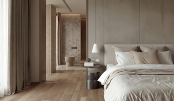 Un dormitorio moderno con una cama grande y sábanas blancas, almohadas de colores neutros, dos lámparas colgantes y mesas de noche. Un baño con paredes de piedra es visible al fondo.