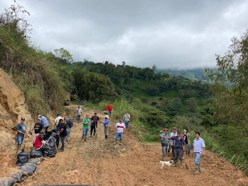 Grupo de personas, la mayoría hombres, en un camino de tierra fangoso en una montaña, usando palas y con sacos negros, rodeados de vegetación y un cielo nublado