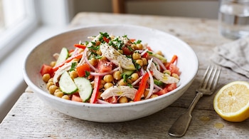 Cuenco blanco con ensalada de garbanzos, pollo, tomate y pepino, junto a medio limón y un tenedor sobre una mesa de madera.