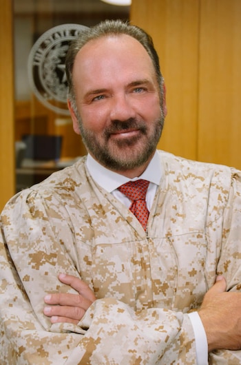 Juez sonriente con barba, vistiendo una toga de camuflaje desierto y corbata roja. Tiene los brazos cruzados y un sello es visible en el fondo
