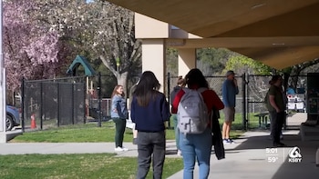 Varias personas, incluyendo estudiantes, paradas bajo un pasillo cubierto en un campus escolar. Al fondo se ve una cerca, un parque infantil y árboles florecidos