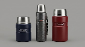 Tres productos Thermos: un termo de comida azul, una botella térmica gris y un termo de comida rojo, con tapas metálicas, sobre fondo gris.