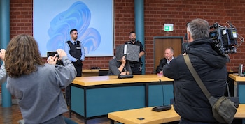 Un hombre en una sala judicial sostiene una carpeta sobre su rostro frente a camarógrafos, agentes de seguridad y un abogado