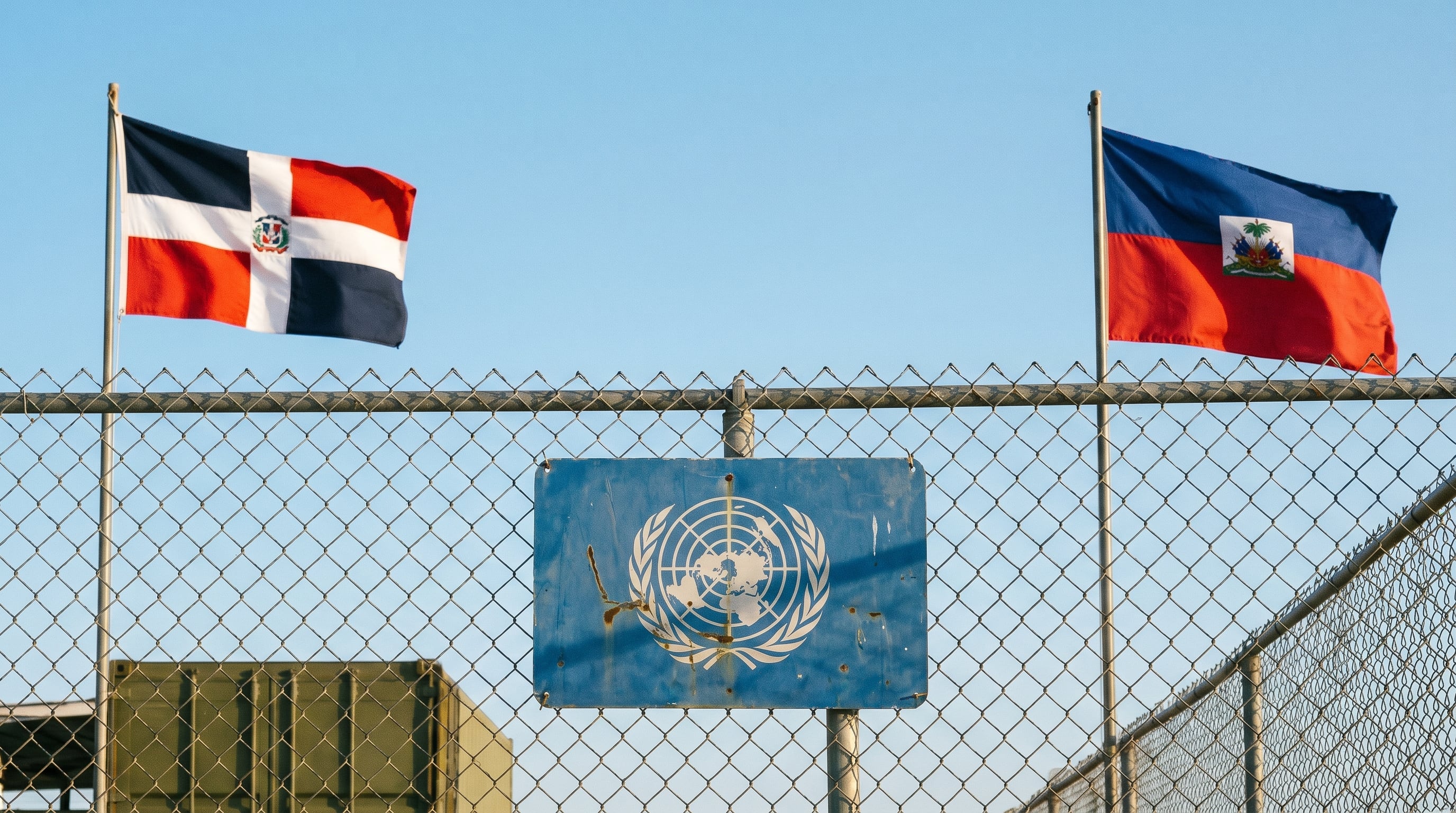 Banderas de República Dominicana y Haití ondean sobre una valla metálica en un punto fronterizo, con un cartel azul de la ONU en el centro, simbolizando la compleja dinámica regional. (Imagen Ilustrativa Infobae)