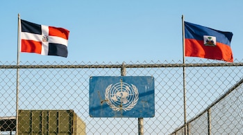 Valla metálica con las banderas de República Dominicana a la izquierda y Haití a la derecha, y un logotipo de la ONU en un cartel azul en el centro, bajo un cielo claro.
