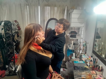 Una persona sentada con cabello largo recibe maquillaje en el rostro. Otra persona le aplica el maquillaje. Hay un tocador con espejo, cosméticos y joyas, y un estante con zapatos