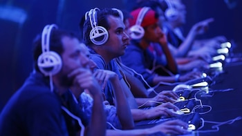 Jugadores sentados en fila con auriculares blancos y ratones, concentrados en pantallas bajo una luz azul brillante en un evento de videojuegos