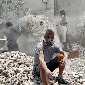Un hombre con mascarilla sentado entre restos de piedra, con dos personas al fondo trabajando con herramientas en un taller