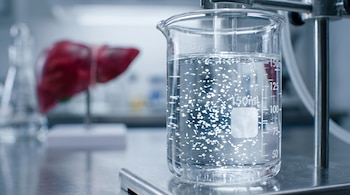 Un vaso de precipitados con partículas brillantes en agua junto a un modelo anatómico de hígado sobre una mesa de laboratorio.