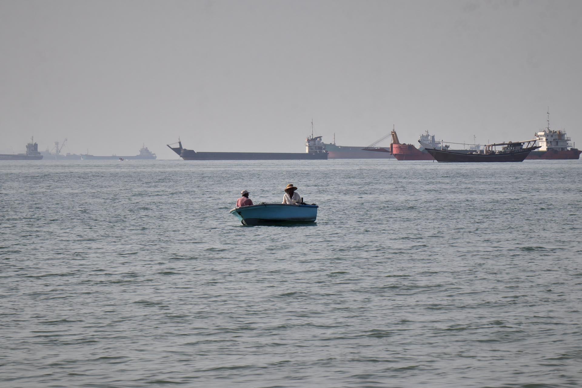 Dos personas navegan en una pequeña embarcación mientras varios buques de carga transitan en las aguas del Golfo, donde un buque fue recientemente capturado frente a las costas de los Emiratos Árabes Unidos y se dirige hacia Irán.