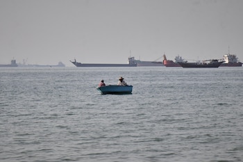 Dos personas en un pequeño bote azul flotan en el mar con ondulaciones ligeras. Al fondo, se ven siluetas de grandes buques de carga bajo un cielo brumoso