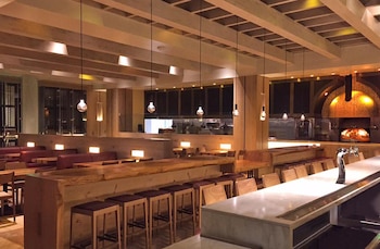 Salón comedor de un restaurante vacío con barras de madera, taburetes altos, mesas, lámparas colgantes y un horno encendido al fondo