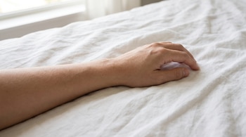Primer plano de un brazo y una mano de persona adulta sobre una sabana blanca arrugada, con una ventana que muestra luz natural al fondo.
