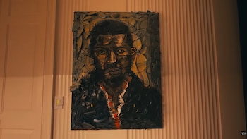 Pintura de un retrato de hombre, tonos amarillos, negros y rojos, con textura. Colgada en una pared acanalada junto a una puerta blanca y un interruptor