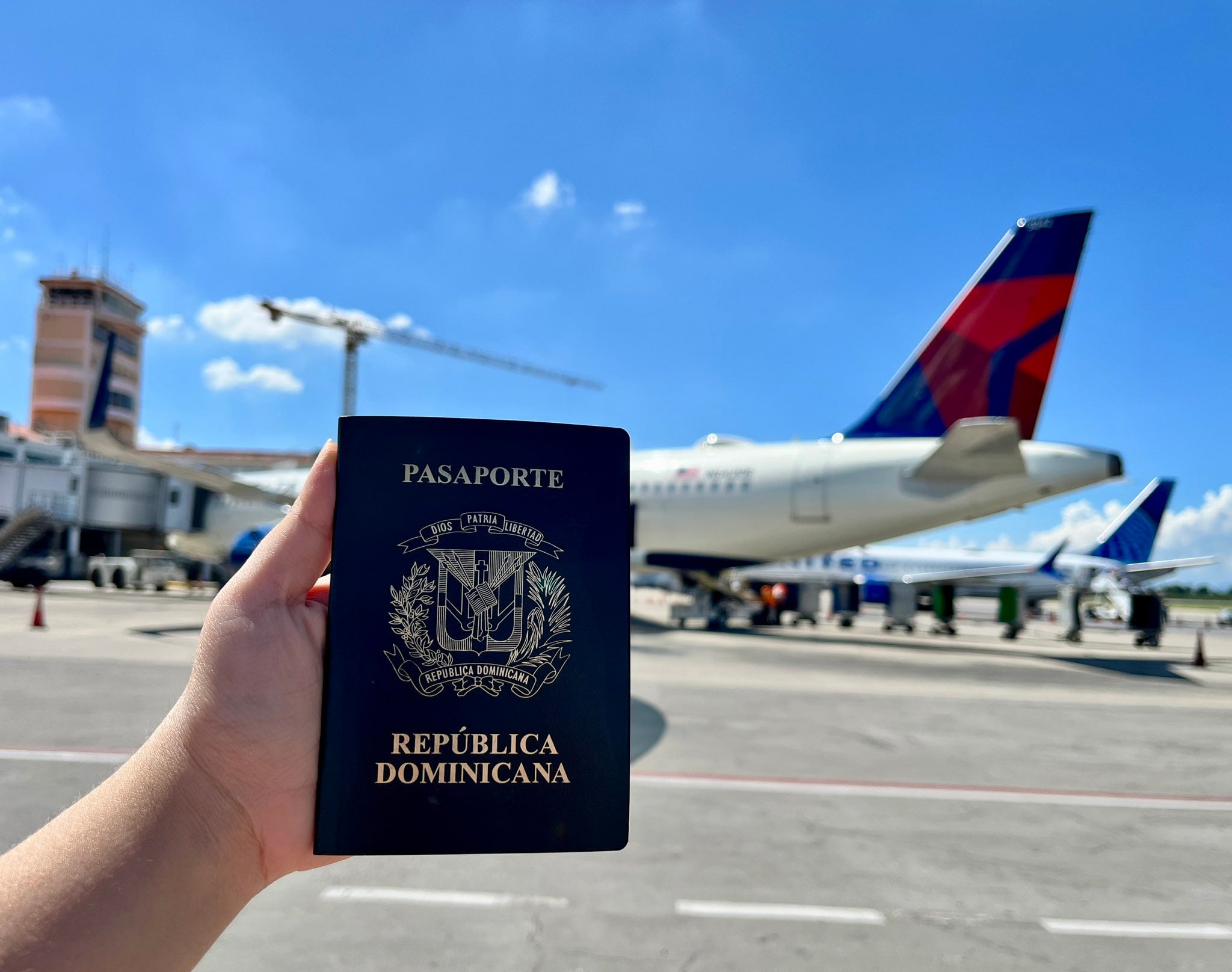 Un pasaporte de la República Dominicana es sostenido en primer plano en la pista del Aeropuerto Internacional del Cibao, con un avión de pasajeros de Delta en el fondo, listo para abordar.