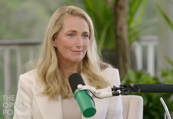 Primer plano de una mujer rubia con cabello ondulado y ojos azules, vistiendo un blazer color crema, sentada frente a un micrófono verde con un fondo borroso de vegetación