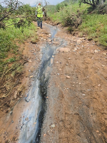 Hombre con chaleco de seguridad camina por un sendero de tierra. Un arroyo con agua grisácea fluye entre la tierra y rocas. Vegetación verde y seca en los márgenes
