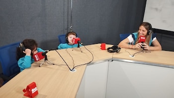 Una mujer y dos niños con auriculares sentados ante una mesa con tres micrófonos de mesa en un estudio con paredes grises