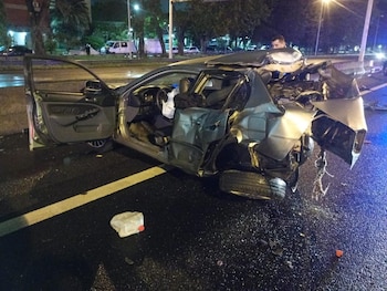 Un automóvil gris con destrozos en la parte trasera y puertas abiertas se encuentra sobre el asfalto mojado de una calle nocturna