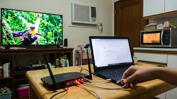 Una mano intenta conectar un cable a una laptop al lado de un router con luces rojas; al fondo, un televisor encendido, un aire acondicionado y un microondas.