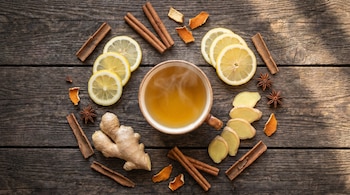 Vista cenital de una taza de infusión dorada humeante en una mesa de madera. Rodeada de rodajas de limón, jengibre fresco, ramas de canela y anís estrellado.