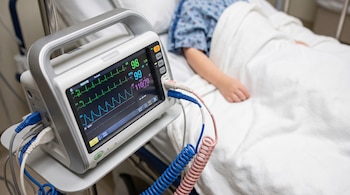 Máquina de monitoreo médico con pantalla que muestra ondas y números, cables conectados y paciente pediátrico cubierto con manta blanca.