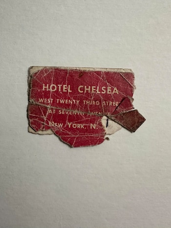 Un fragmento de papel rojo, viejo y rasgado, con el texto "HOTEL CHELSEA WEST TWENTY THIRD STREET AT SEVENTH AVENUE NEW YORK, N." visible y descolorido