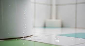 Primer plano de gotas de líquido transparente, presumiblemente vinagre blanco, goteando por la base de un inodoro blanco y sobre azulejos claros con secciones verdes y azules.