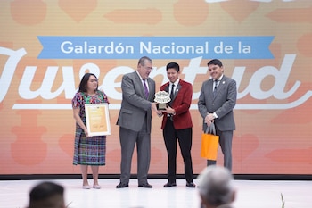 Cuatro personas en un escenario. Un hombre entrega un trofeo a un joven. Una mujer sostiene un certificado. Una pantalla de fondo dice 'Galardón Nacional de la Juventud'