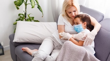 Una mujer y una niña con mascarilla médica azul y guantes se abrazan sentadas en un sofá