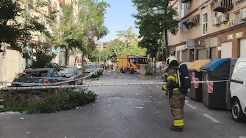 Un bombero de espalda con casco y equipo, calle con escombros, árboles caídos, dos coches dañados, un camión de bomberos, cinta de "NO PASAR" y contenedores de basura