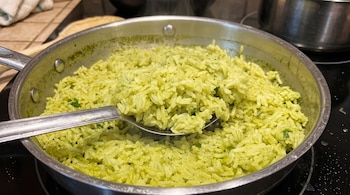 Sartén de acero inoxidable sobre una cocina con arroz de tono verde y una cuchara metálica que sostiene una porción del grano.