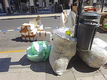 Una acera llena de varias bolsas de basura blancas y verdes, cajas de cartón atadas y un bote de basura gris desbordado junto a una carretera