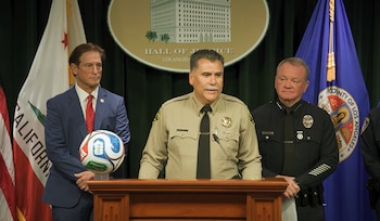 Tres hombres, incluyendo el sheriff Robert Luna, en uniformes y traje formal en un podio con banderas de California y Los Ángeles. Un balón de fútbol está a la izquierda