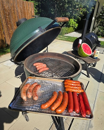 Una parrilla verde abierta con salchichas en la rejilla, una bandeja llena de embutidos y media sandía al fondo