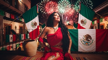 Mujer con vestido rojo y collar turquesa en un patio rodeada por banderas de México, papel picado y fuegos artificiales nocturnos.