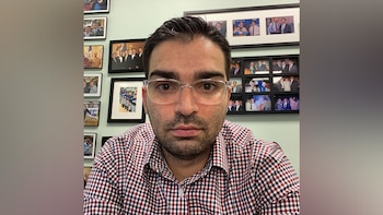 Hombre con lentes y camisa a cuadros frente a una pared verde con múltiples fotos enmarcadas
