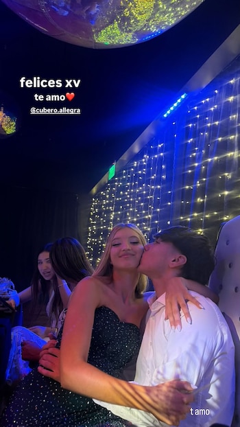 Lucho Ferrari besa la mejilla de Allegra Cubero. La joven usa vestido oscuro, el joven camisa blanca. Luces y personas en el fondo. Texto 'felices xv te amo'