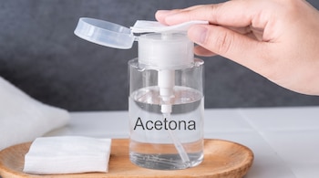 Botella transparente con la palabra Acetona, una mano que presiona un disco de algodón en el dispensador y una bandeja de madera.