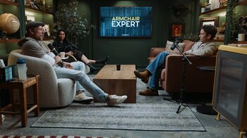 Tres personas sentadas en un estudio de podcast con muebles modernos; dos hombres y una mujer. Hay micrófonos, una mesa de madera y una pantalla que muestra 'Armchair Expert'