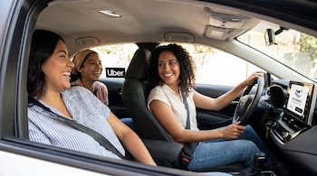 Interior de un auto Uber con tres mujeres sonrientes. Una mujer conduce y dos son pasajeras. En la pantalla del auto se ve "Uber Mujeres".
