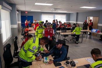 Salón de capacitación con varias personas en uniformes de emergencia, una de ellas mide la presión arterial a otra persona con tensiómetro y estetoscopio
