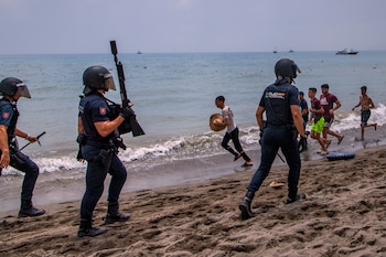 Tres agentes de policía con cascos y equipamiento táctico en una playa. Varias personas corren por la orilla y el agua. Barcos en el mar y cielo gris