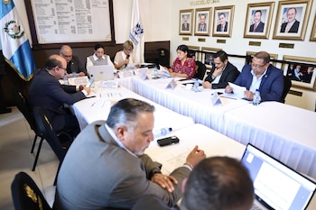 Personas reunidas alrededor de una mesa con documentos y computadoras portátiles frente a la bandera de Guatemala